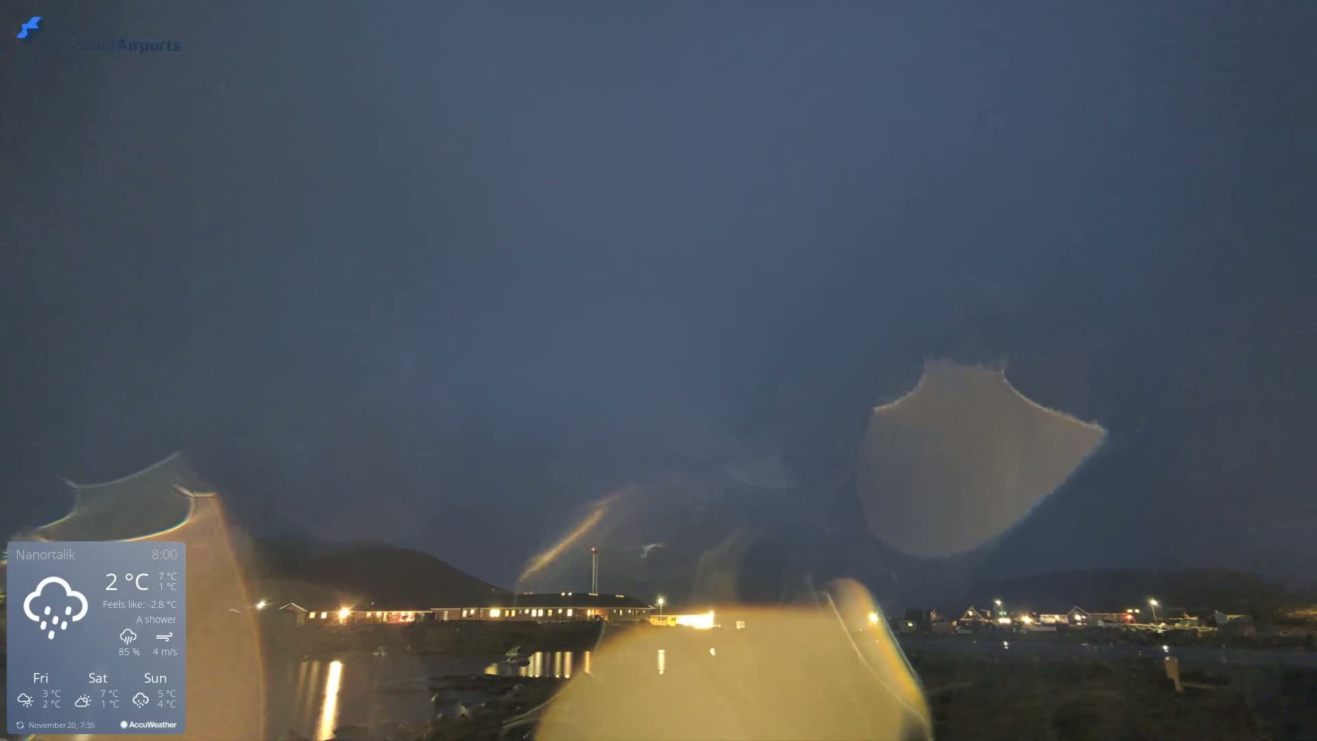 A dark, overcast, and rainy outdoor scene captured through a wet lens shows artificial lights from buildings reflecting on a body of water, with hills visible in the distant background.