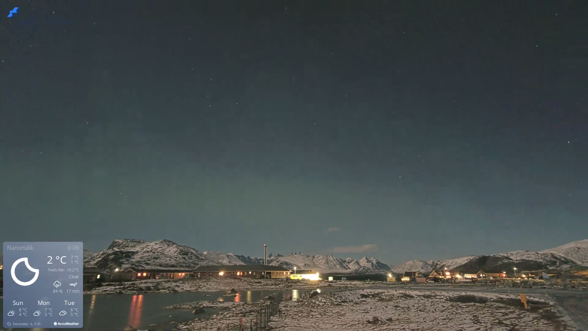A clear, snowy night scene displays an illuminated village nestled among dark mountains, with lights reflecting in a body of water, all beneath a starry sky exhibiting a subtle green auroral glow.