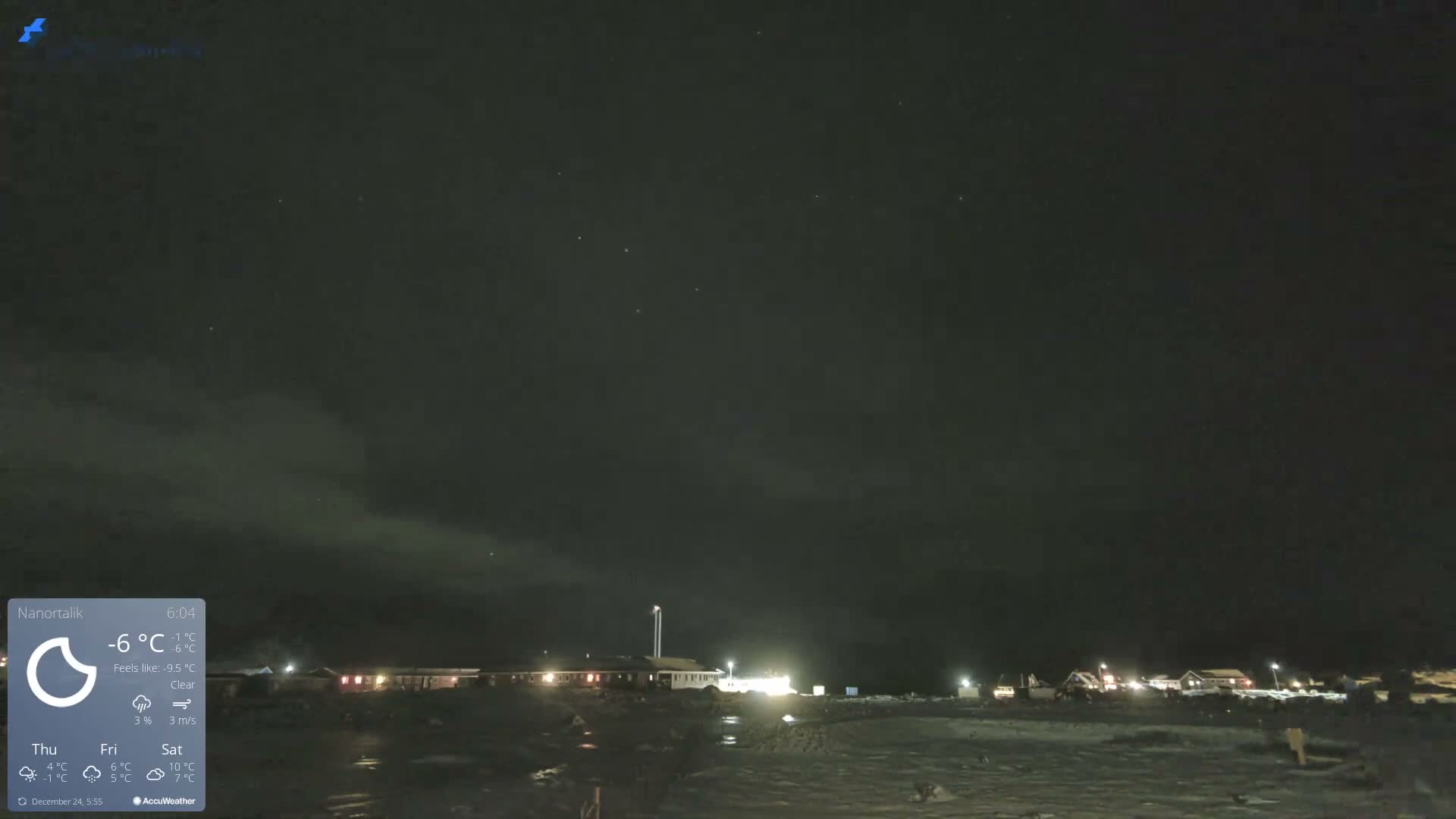 An outdoor scene at night shows illuminated buildings on snow-covered ground under a dark, clear sky with faint visible stars.