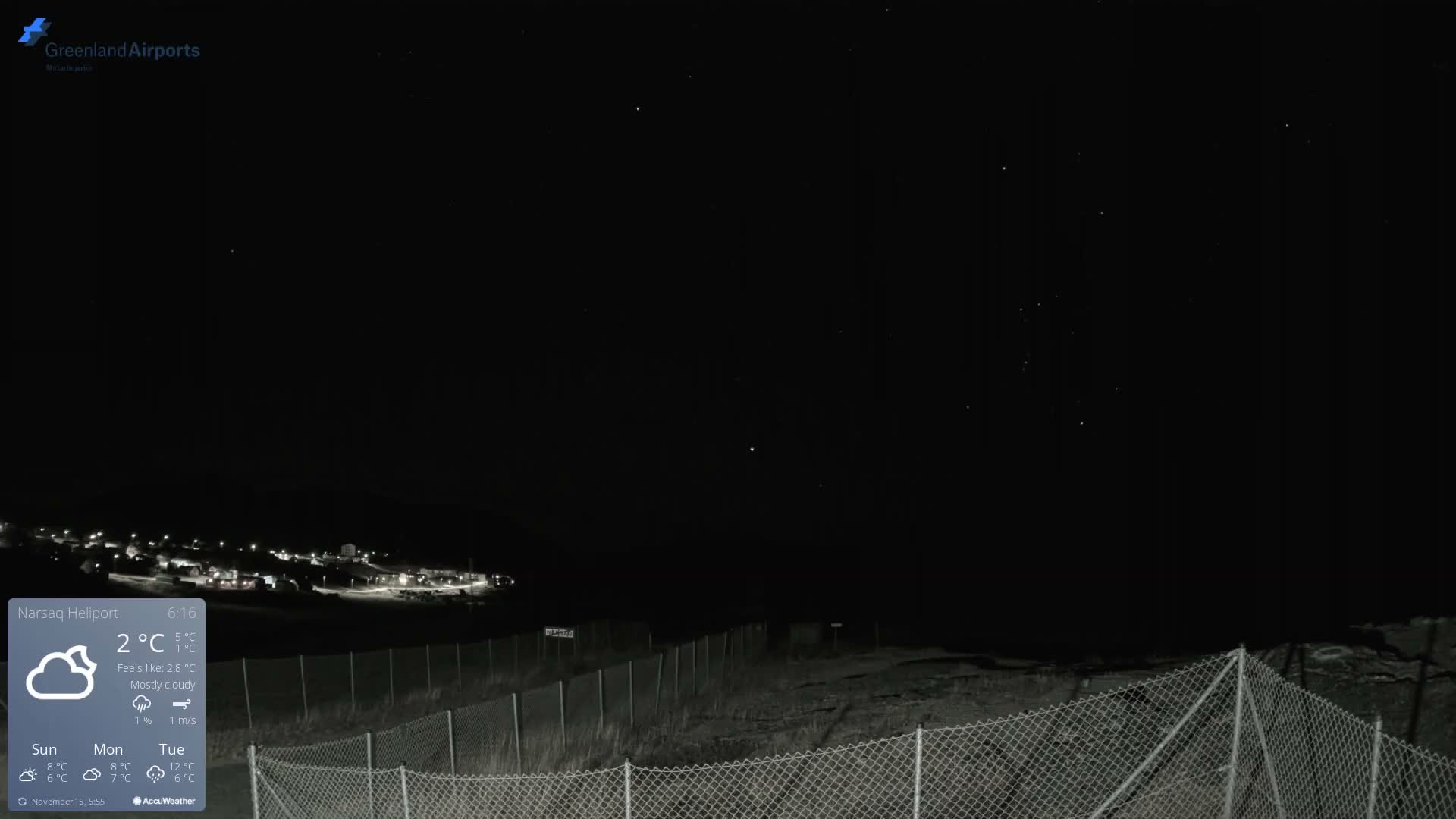 A dark, clear night scene shows a chain-link fence in the foreground, a dimly lit grassy area, and a distant settlement with scattered lights against a star-speckled sky.
