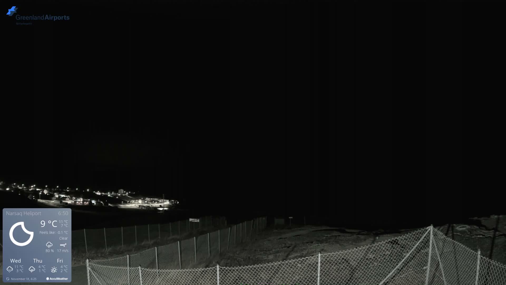 A dark, clear night view shows a chain-link fence in the foreground with the distant lights of a settlement visible against the dark horizon.