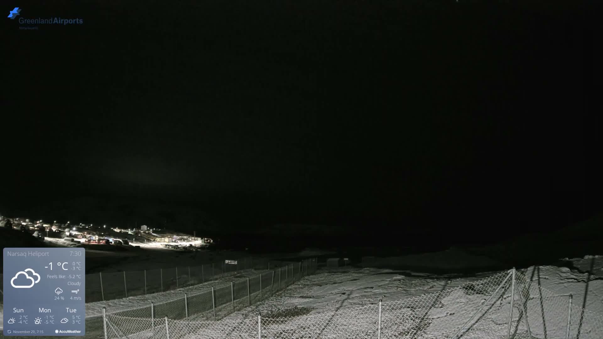 A snowy, cloudy night scene features a chain-link fence in the foreground, leading towards a distant settlement with scattered lights visible against the dark, overcast sky.