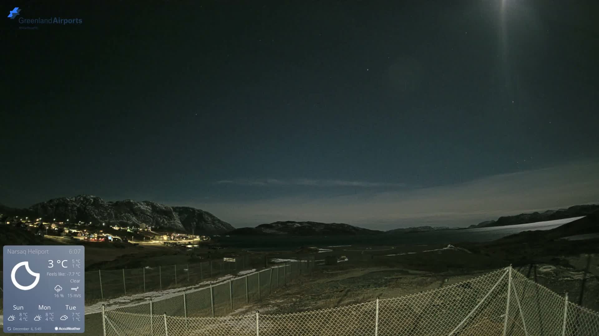 A clear night sky with visible stars and a prominent moonbeam illuminates a landscape featuring a small, lit-up settlement nestled among snow-capped mountains, a body of water, and a snowy field with a chain-link fence in the foreground.