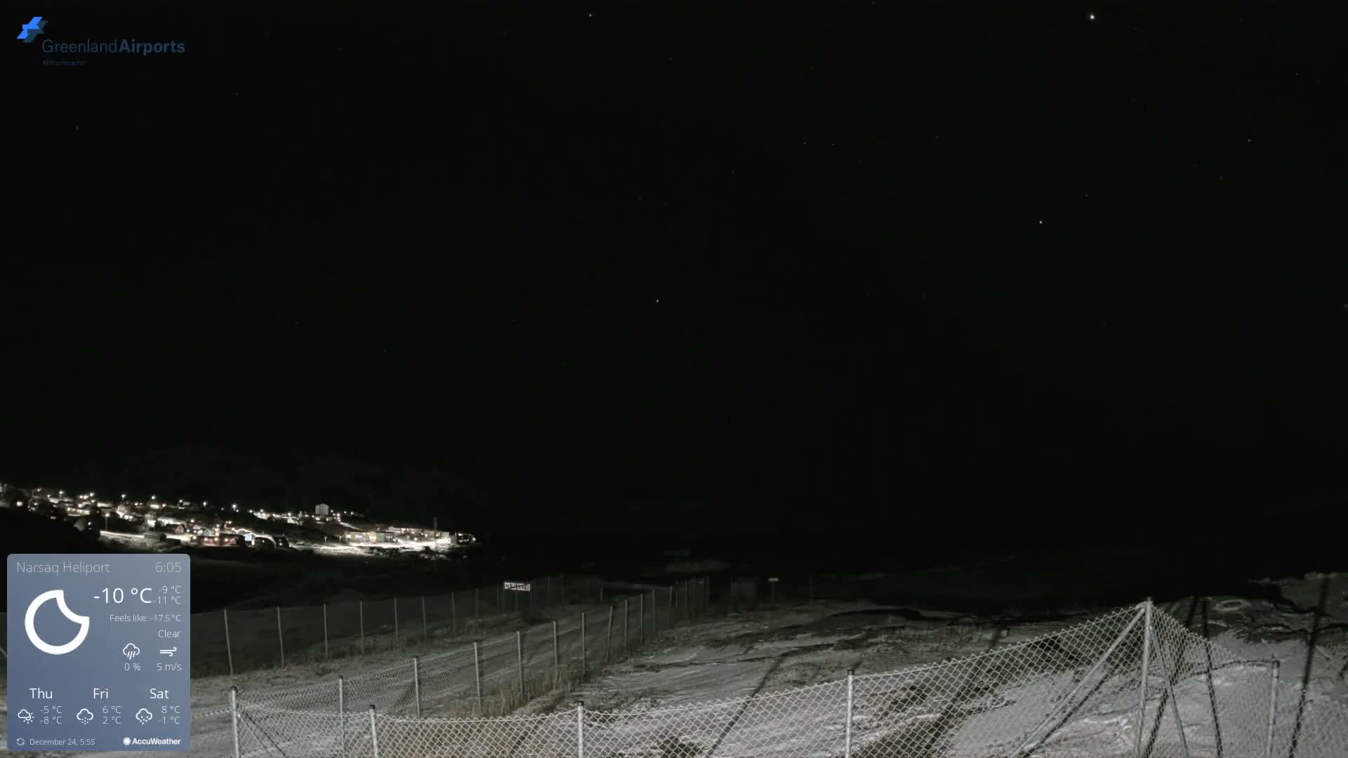 A clear, cold night reveals a snow-covered landscape with a brightly lit town on a distant hillside under a star-filled sky, viewed across a chain-link fence in the foreground.
