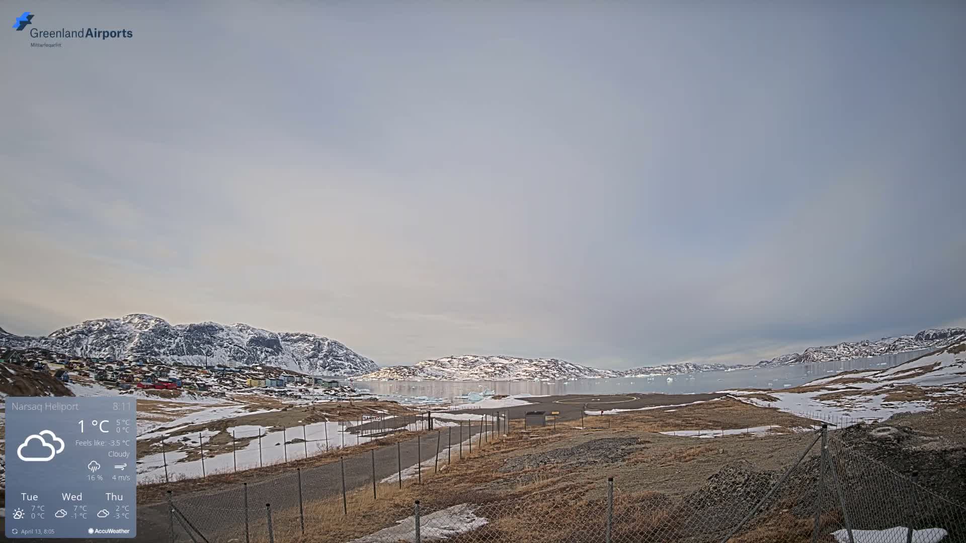 A clear, cold night reveals a snow-covered landscape with a brightly lit town on a distant hillside under a star-filled sky, viewed across a chain-link fence in the foreground.