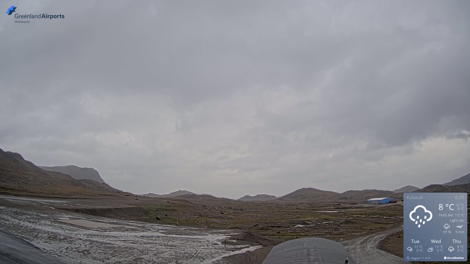 A flat, barren landscape with low, dark-colored hills under a gray, overcast sky experiences light rain.