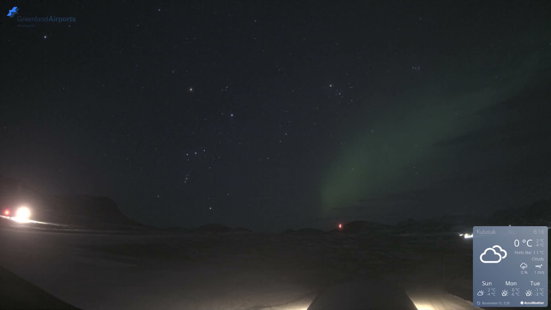A clear, star-filled night sky, displaying a vibrant green aurora borealis, illuminates a snow-covered landscape punctuated by bright artificial lights and distant hills.