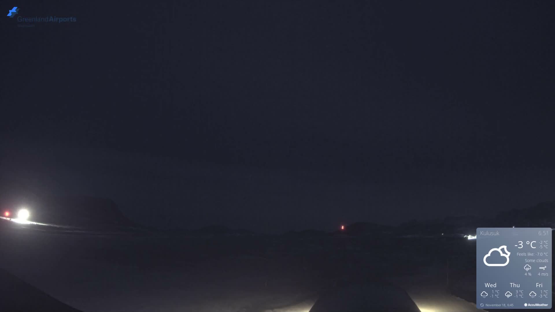 A dark, snow-covered outdoor landscape at night is illuminated by bright lights on the left and a distant red beacon on a hill, all under a heavily overcast or dark sky, suggesting cold conditions.