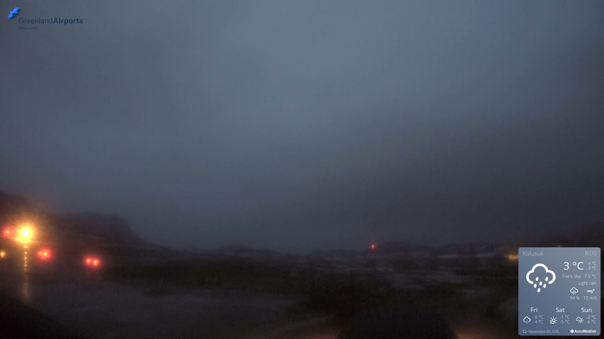 A dark and blurry outdoor scene reveals an indistinct, rugged landscape with bright yellow and red lights on the left, under conditions of low visibility and light rain.