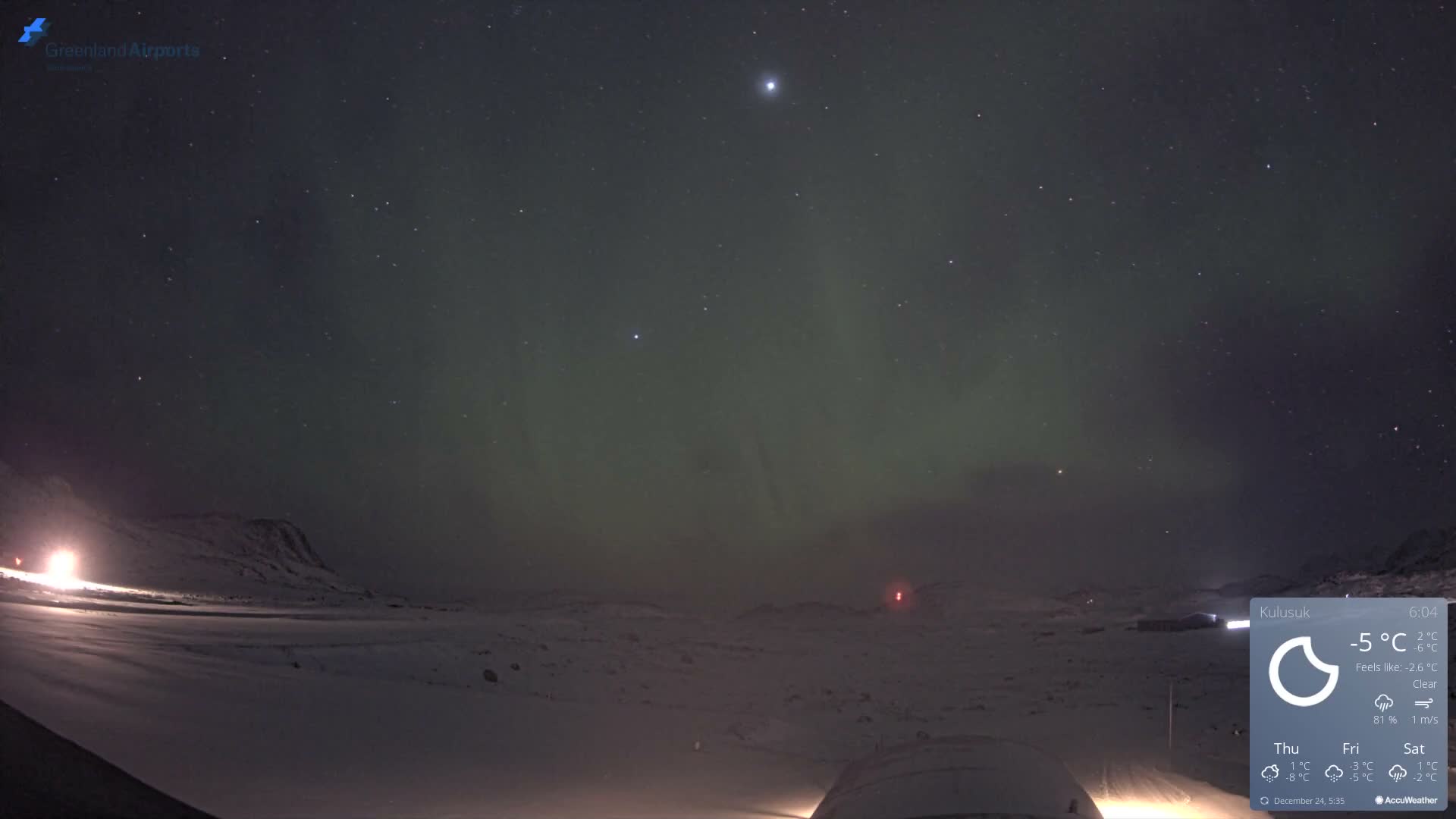 A dark, clear night sky displaying numerous stars and a green aurora borealis hangs over a vast snow-covered landscape with distant mountains and a few ground lights, suggesting cold and clear weather.