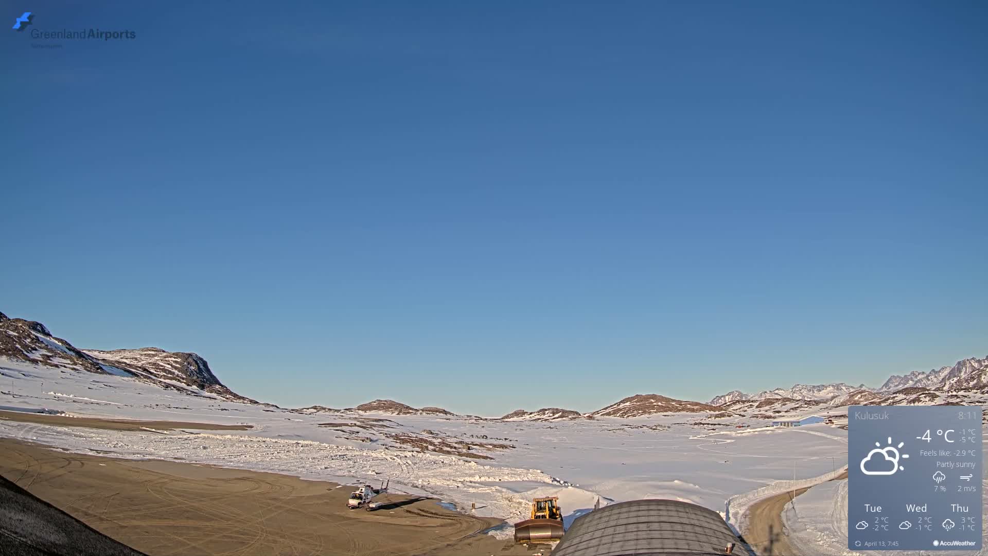 A dark, clear night sky displaying numerous stars and a green aurora borealis hangs over a vast snow-covered landscape with distant mountains and a few ground lights, suggesting cold and clear weather.