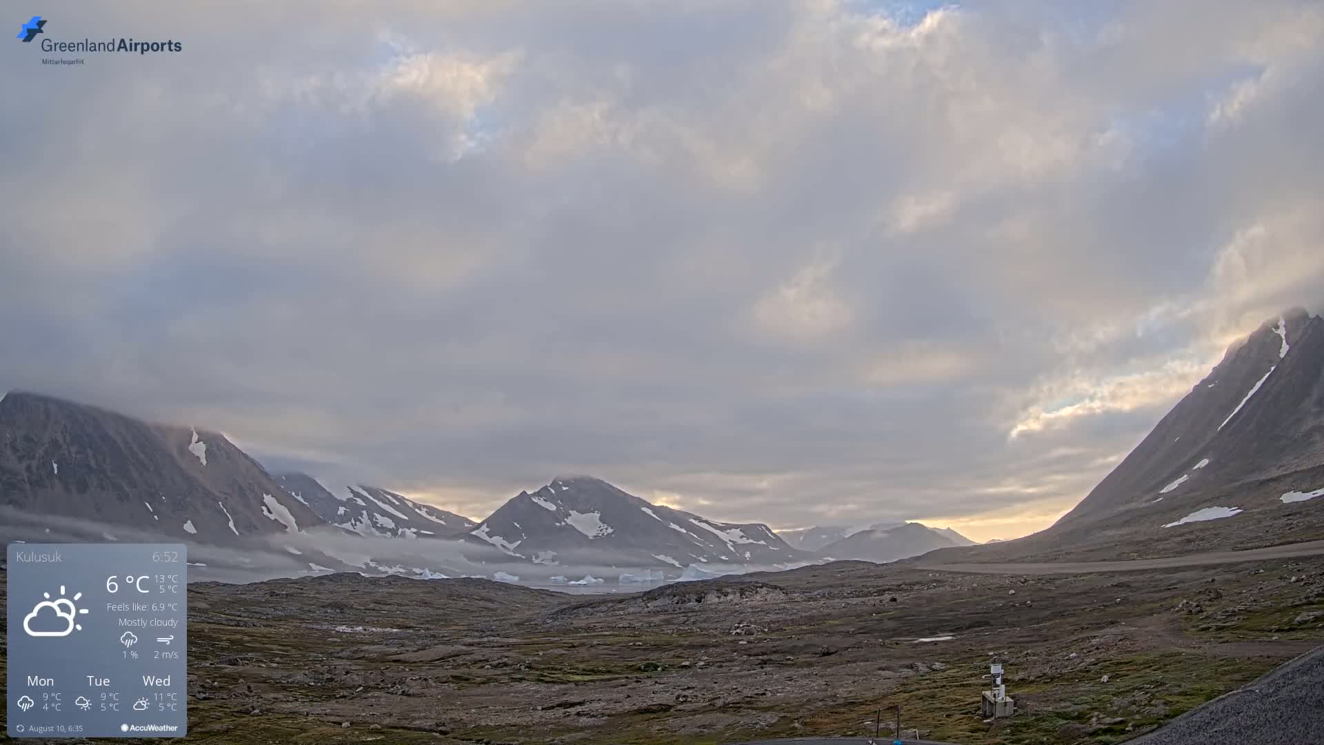 A mostly cloudy, cool landscape features snow-capped mountains and a flat, dark-colored plain with patches of vegetation under a mostly cloudy sky.