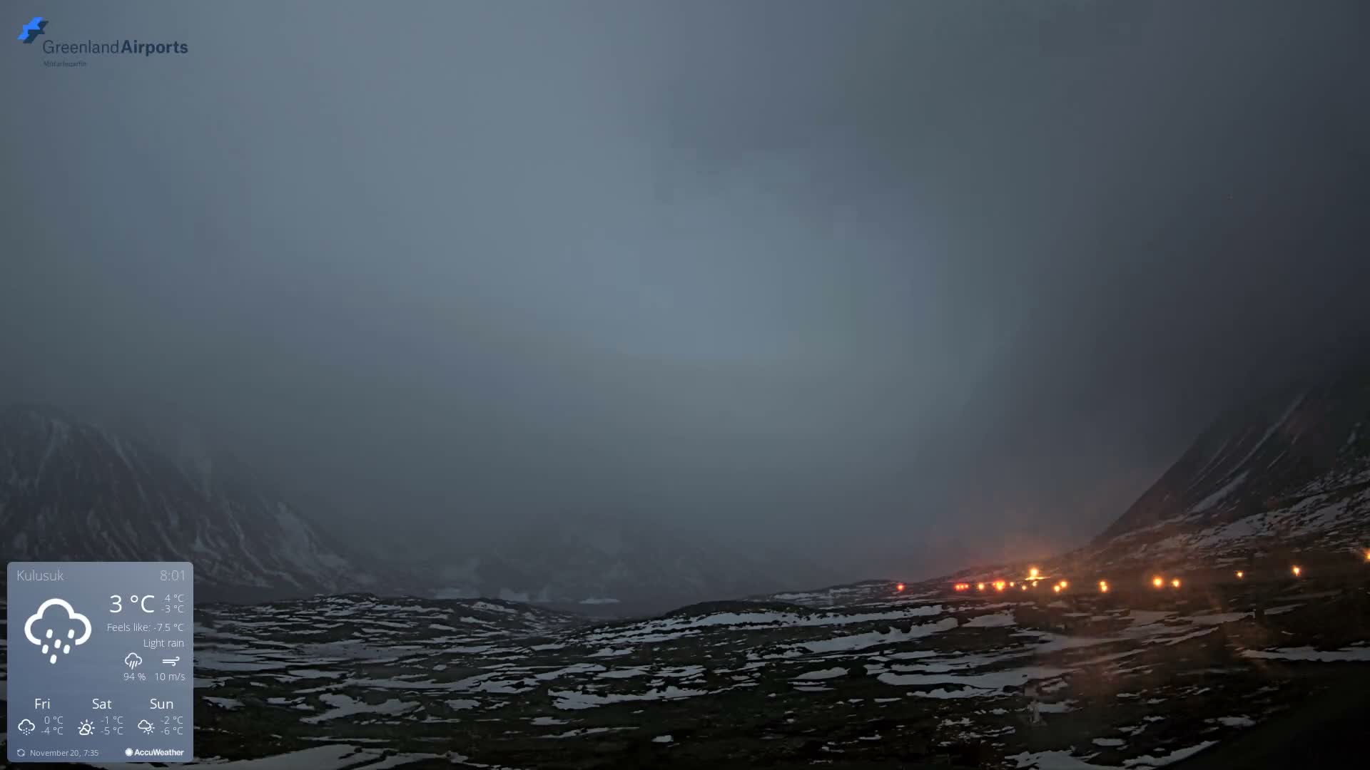 A vast, snow-dusted, rocky landscape with mountains shrouded in fog and light rain is seen under dim, overcast conditions, with a line of bright orange lights stretching along the right side of the view.
