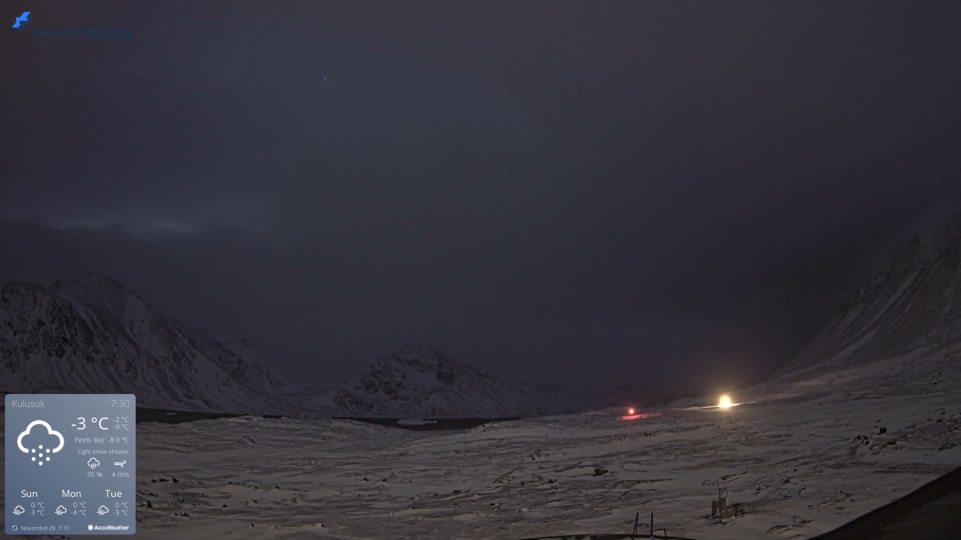 A vast, snow-covered mountainous landscape appears dark under an overcast sky at night or early morning, with distant bright lights illuminating a cold, snowy scene.