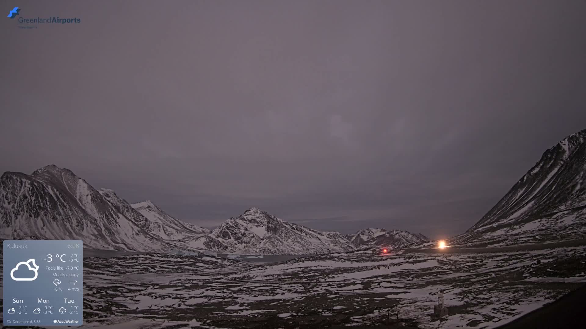 A vast, desolate, snow-covered mountainous landscape is visible under a dim, mostly cloudy sky, with bright artificial lights illuminating a distant area.