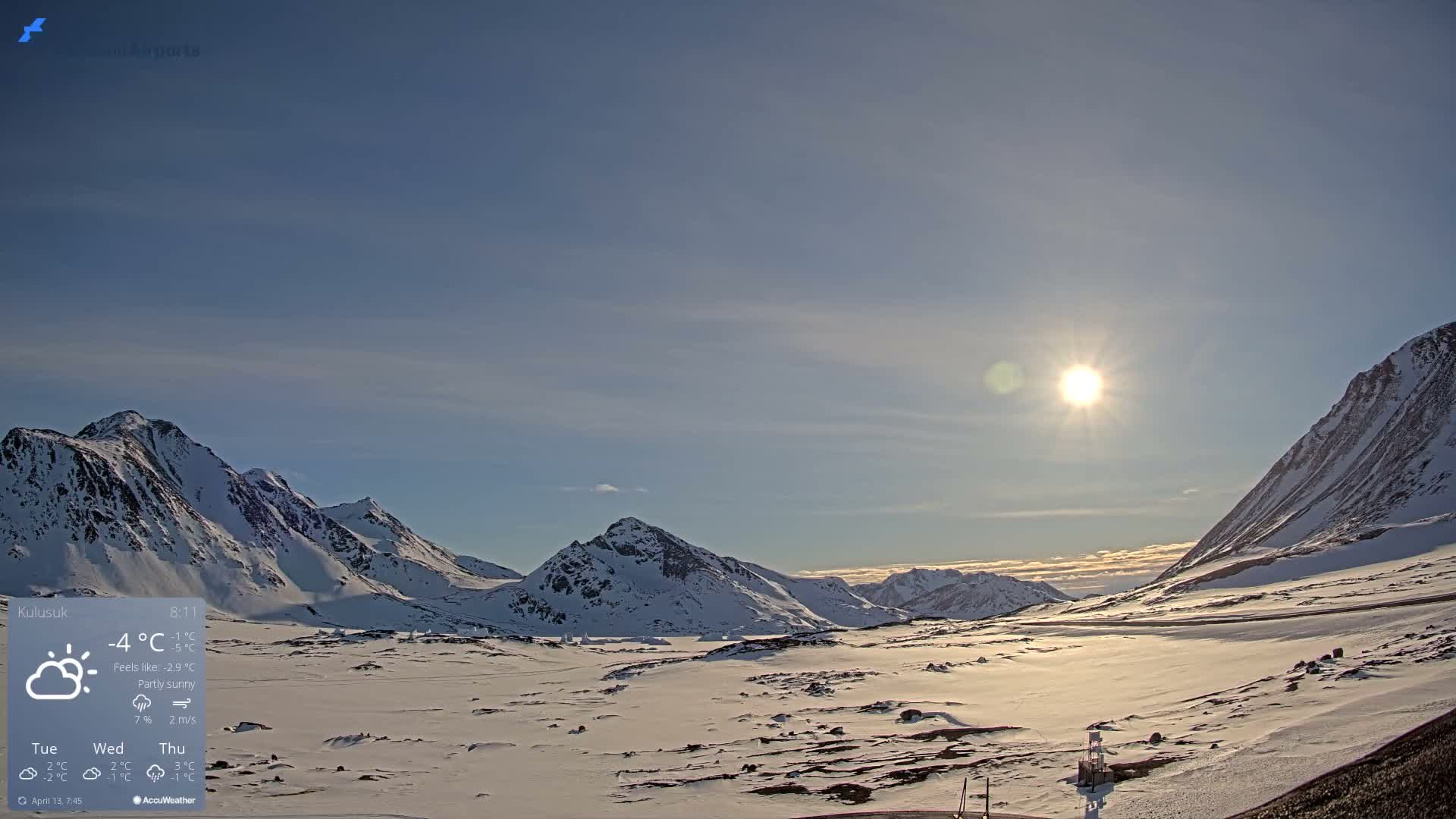 A vast, clear, snow-covered mountainous landscape is illuminated by a starry night sky with faint green aurora borealis, featuring distant lights across the snowy ground.