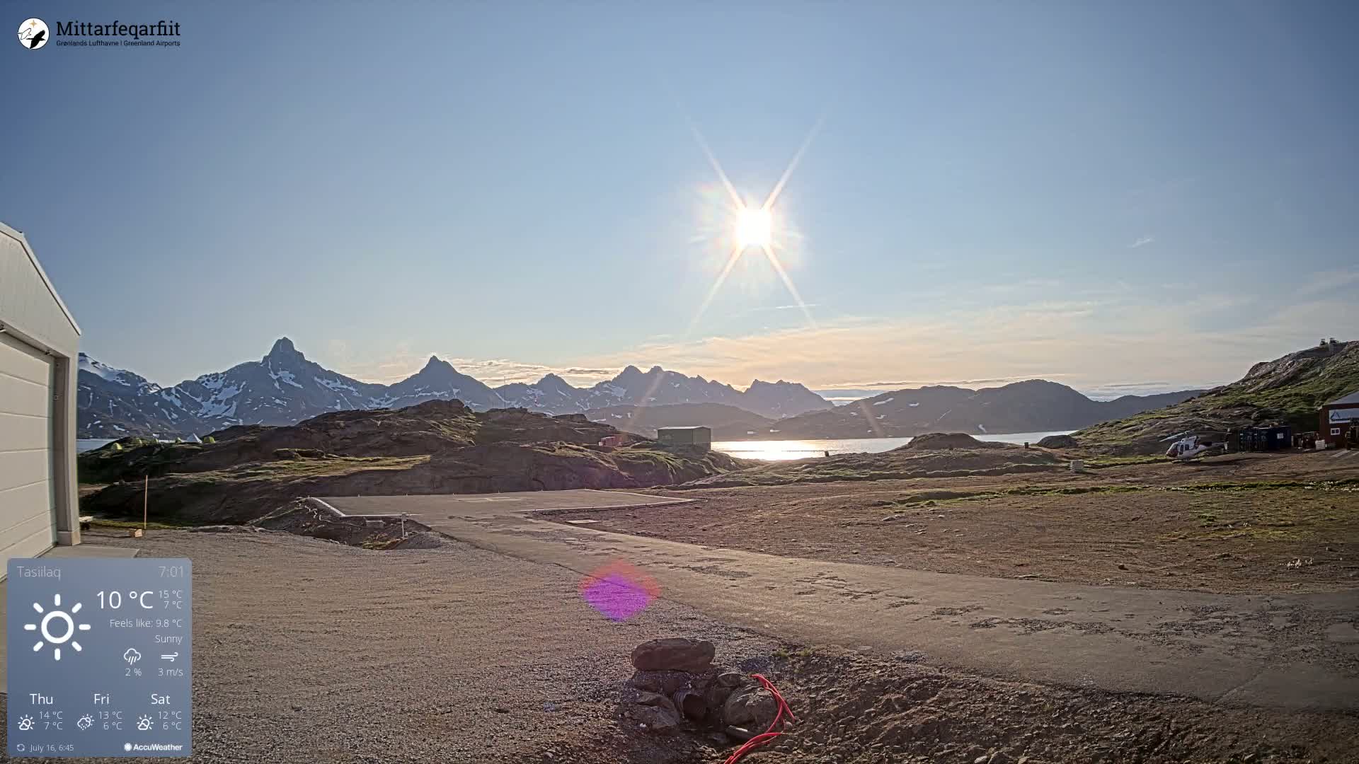 A sunny day reveals a flat, rocky landscape near a body of water, with a helicopter visible in the distance and mountains in the background.