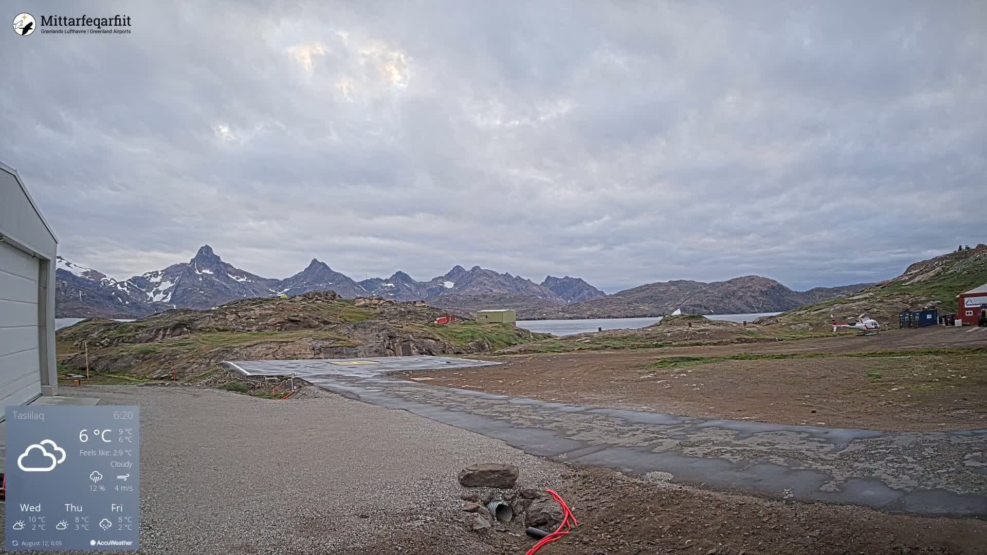 A helicopter sits on a helipad near a building, overlooking a rocky coastline and a range of snow-capped mountains under a cloudy sky.