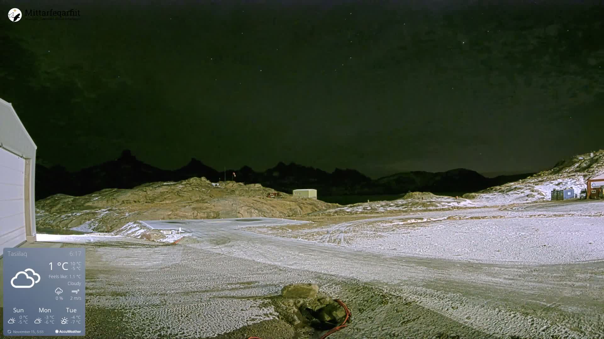 A dark, starry night blankets a rugged, snow-dusted landscape with distant mountains and scattered small buildings.