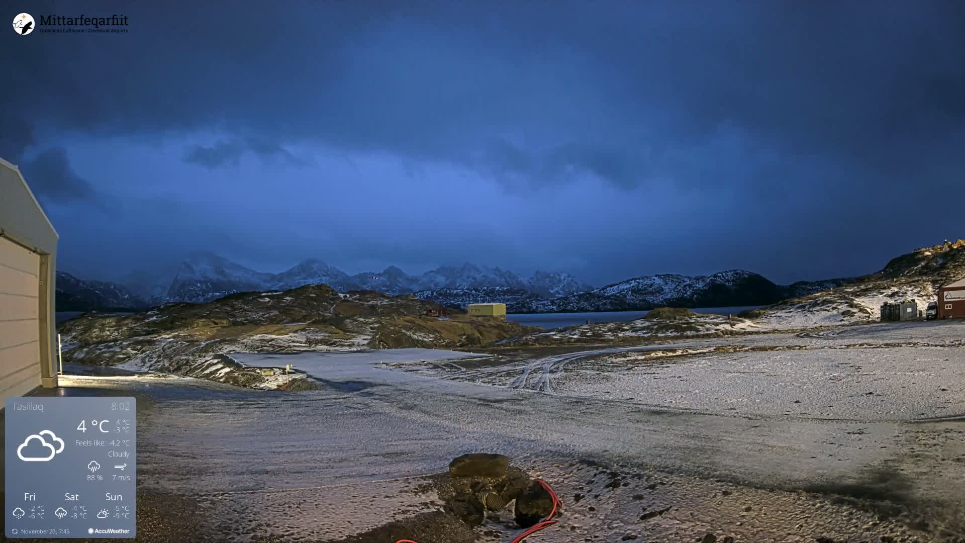 A wide, wintry landscape features snow-dusted, rugged terrain with distant mountains, a calm body of water, and a few scattered buildings under a dark, heavily overcast sky, indicating cold and cloudy outdoor conditions.