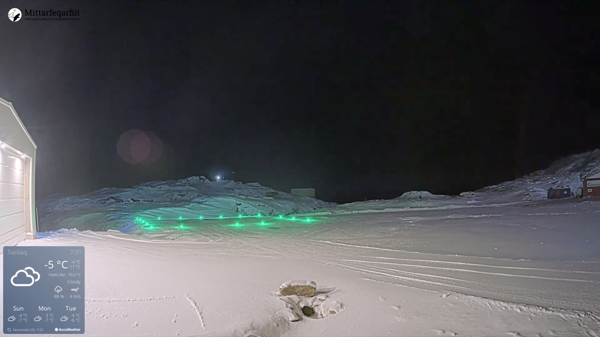 A snowy outdoor landscape at night features green runway lights illuminating a vast snow-covered ground with distant hills and buildings, all under cloudy skies with a temperature of -5°C.