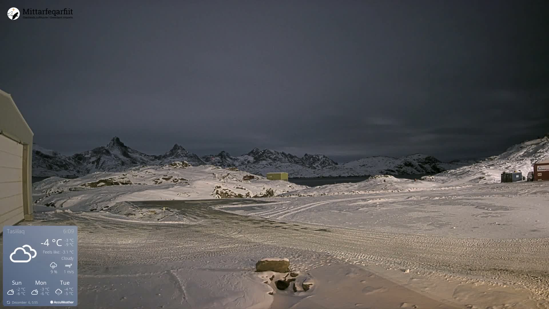 A stark, snow-covered landscape stretches under a dark, cloudy sky, revealing rugged, snow-capped mountains, a body of water, and scattered small structures across the wintry terrain.