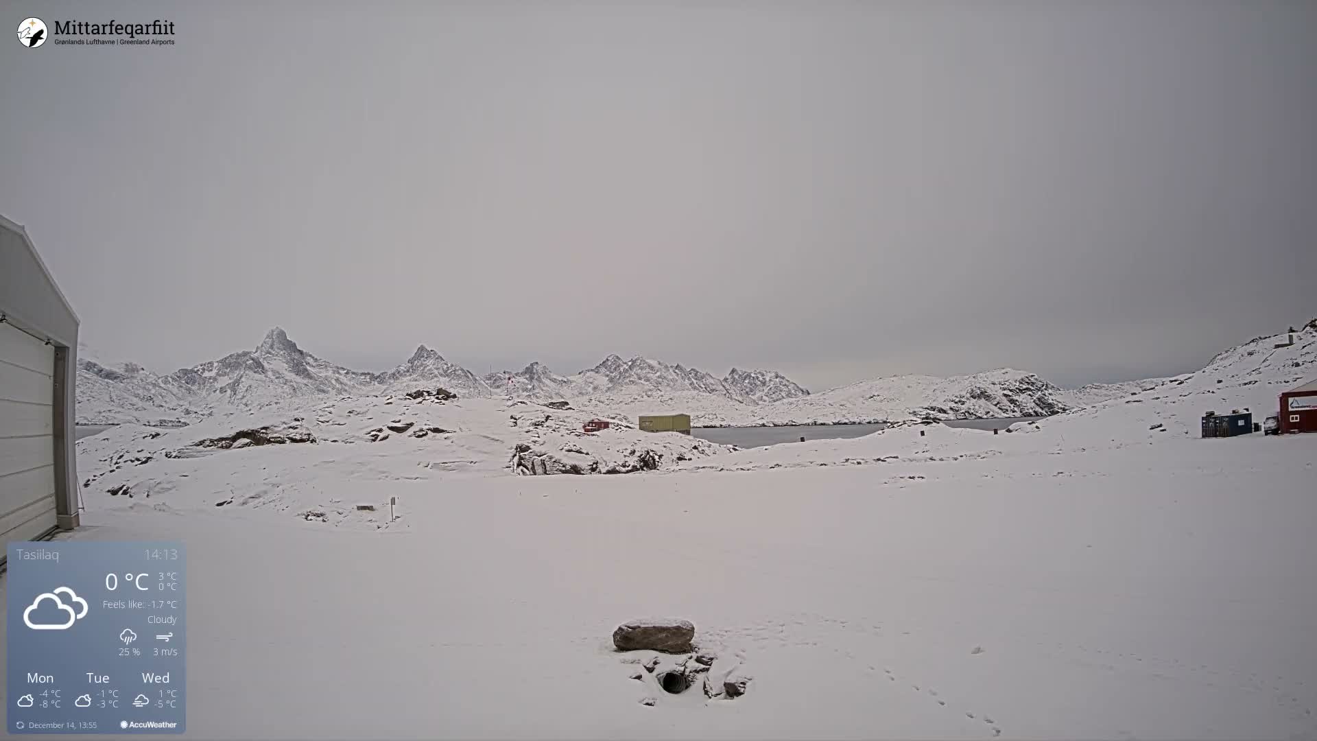 A stark, snow-covered landscape stretches under a dark, cloudy sky, revealing rugged, snow-capped mountains, a body of water, and scattered small structures across the wintry terrain.