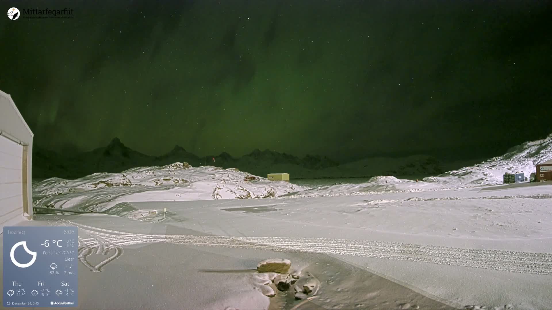 A clear and very cold winter night reveals a vibrant green aurora borealis dancing above snow-covered mountains and terrain, with tire tracks and a heart etched into the fresh snow in the foreground.