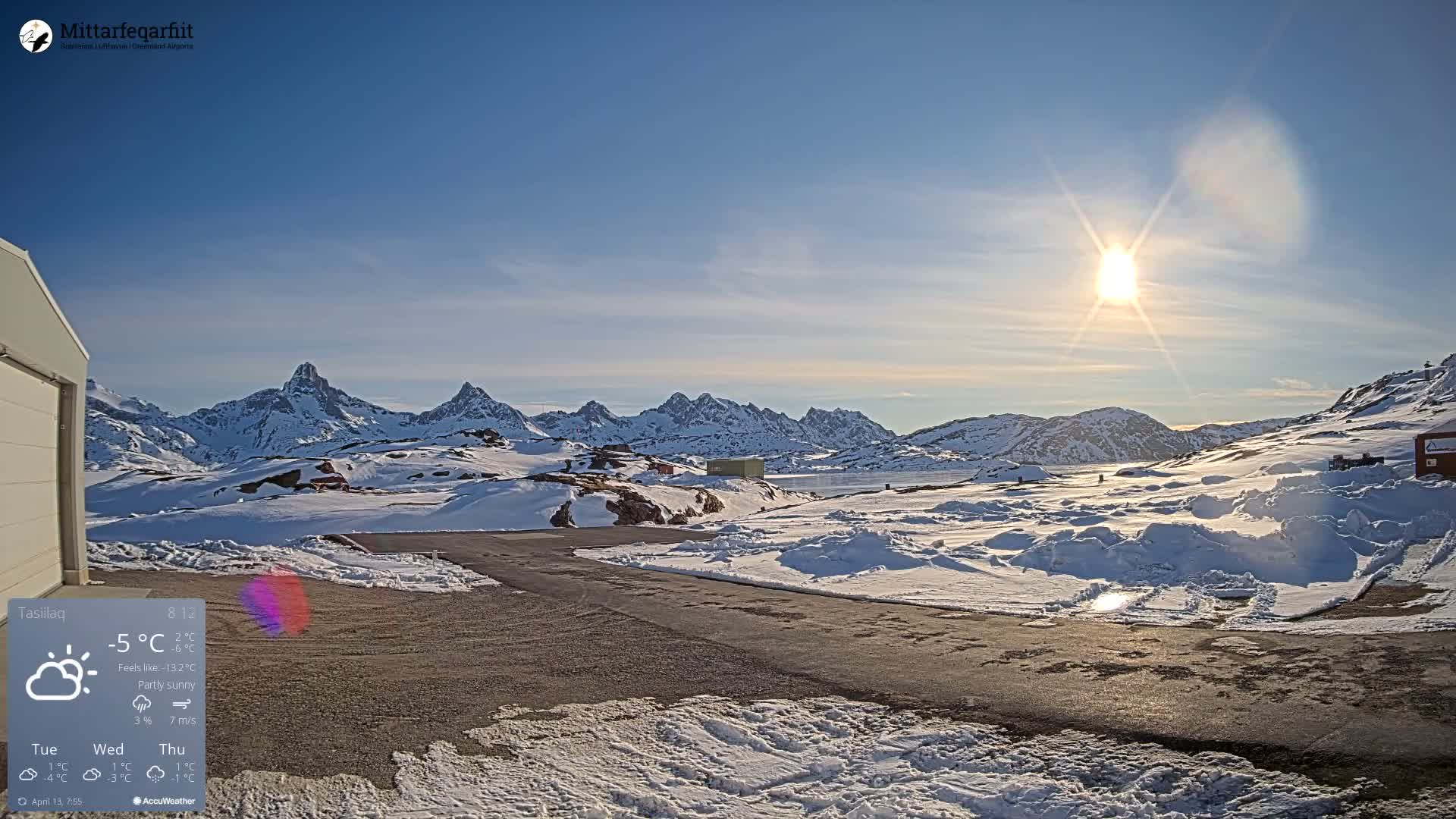 A clear and very cold winter night reveals a vibrant green aurora borealis dancing above snow-covered mountains and terrain, with tire tracks and a heart etched into the fresh snow in the foreground.