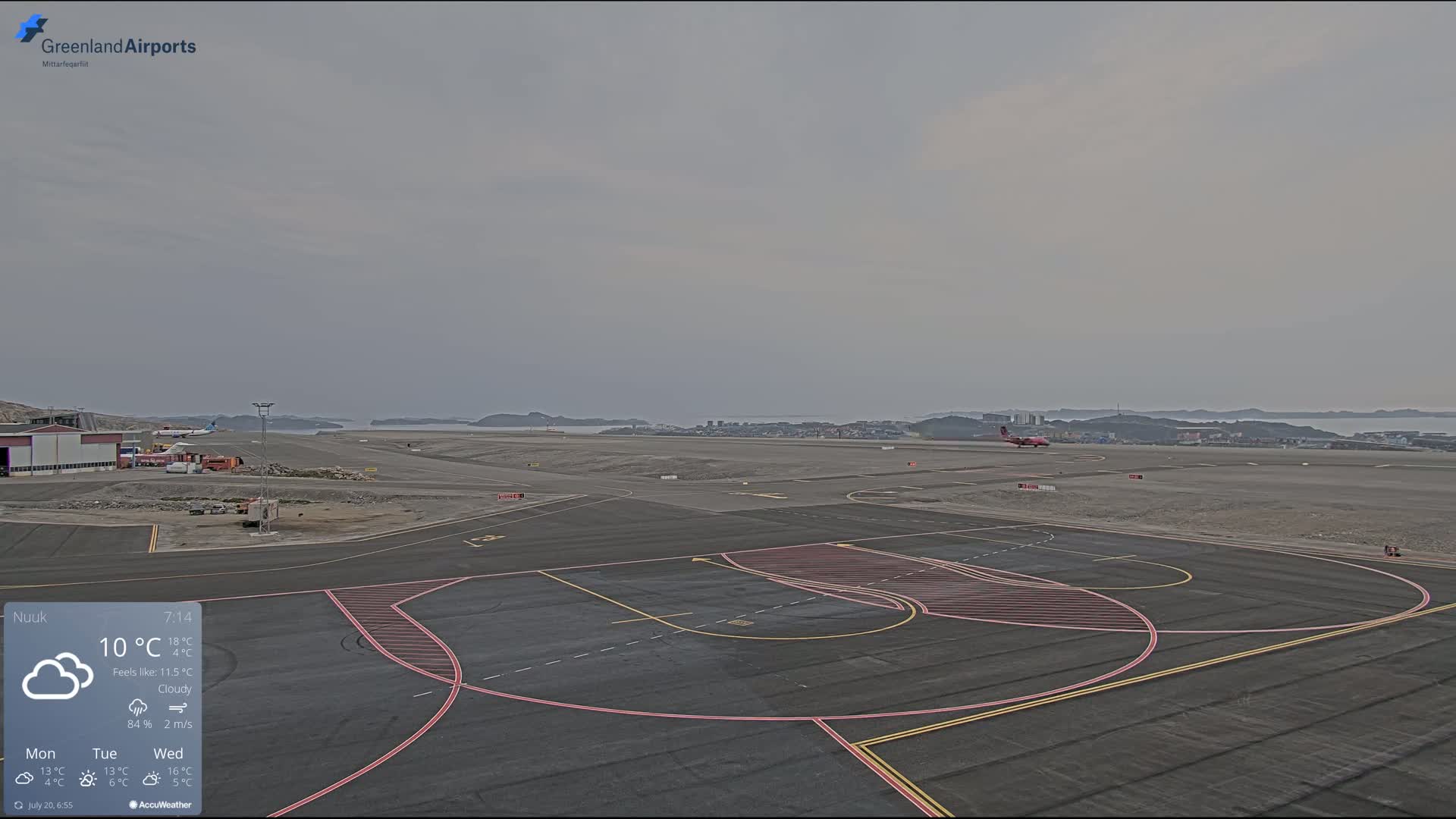 A mostly cloudy airport tarmac is shown with a small airplane on the runway and a distant town visible across the water.