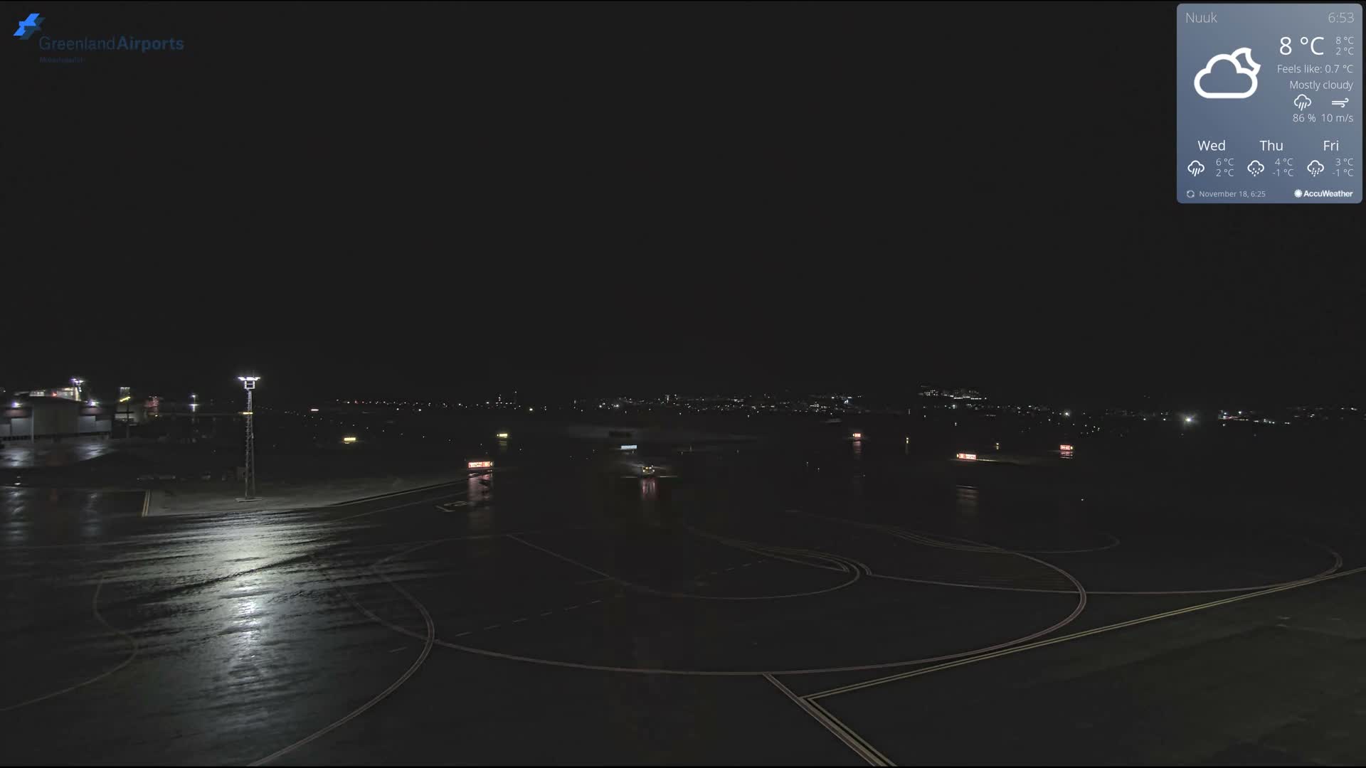 A wet airport tarmac, illuminated by sparse artificial lights reflecting on its surface, extends towards distant city lights under a dark, nighttime sky, suggesting recent rain or damp conditions.