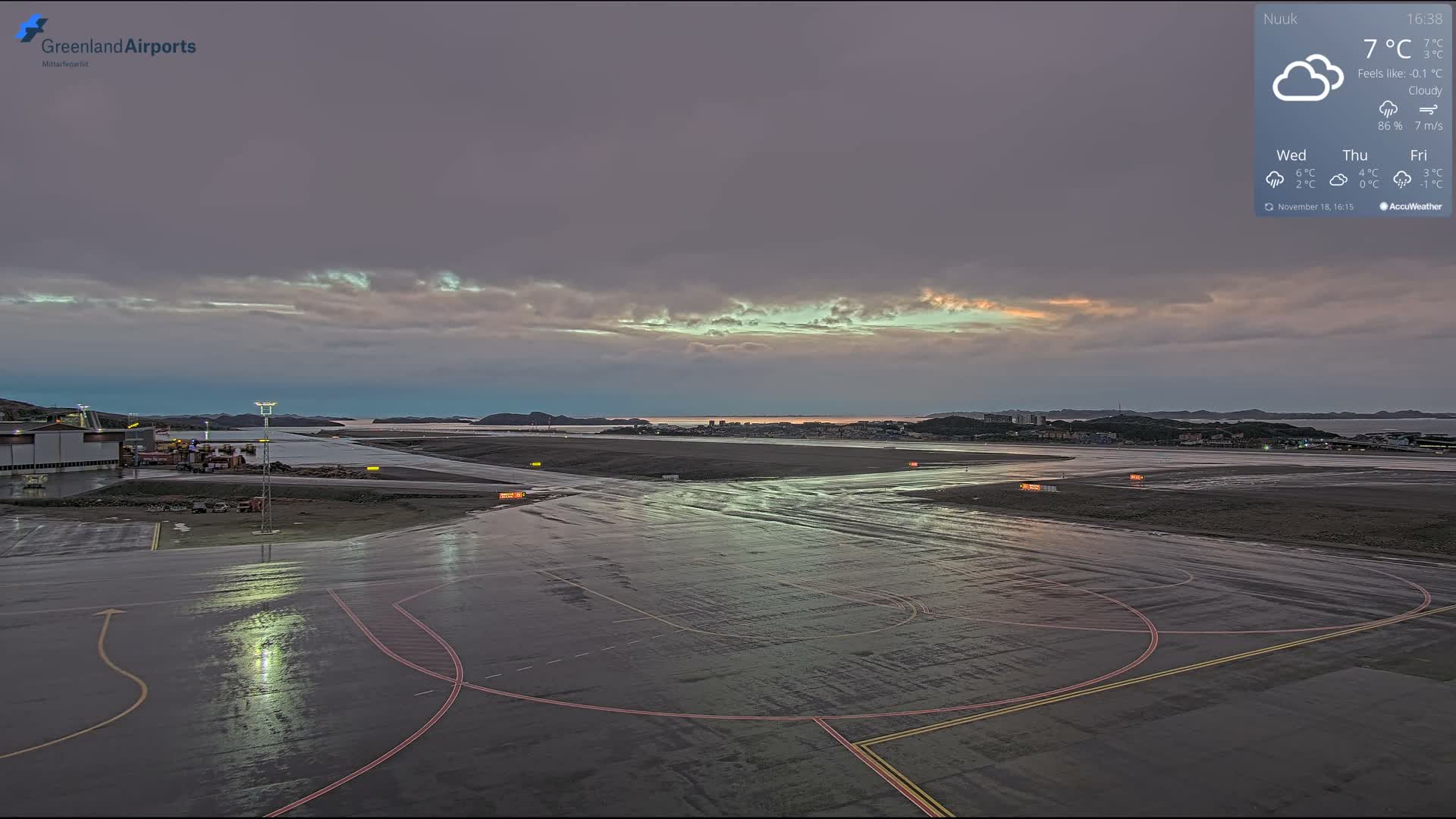 A wet airport tarmac, illuminated by sparse artificial lights reflecting on its surface, extends towards distant city lights under a dark, nighttime sky, suggesting recent rain or damp conditions.