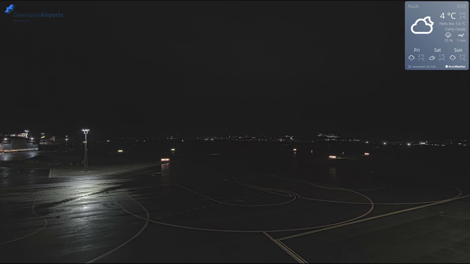 The image displays a wet airport tarmac and taxiway, illuminated by an overhead light pole and various airport lights, with distant city lights visible under a partly cloudy night sky at 4°C.