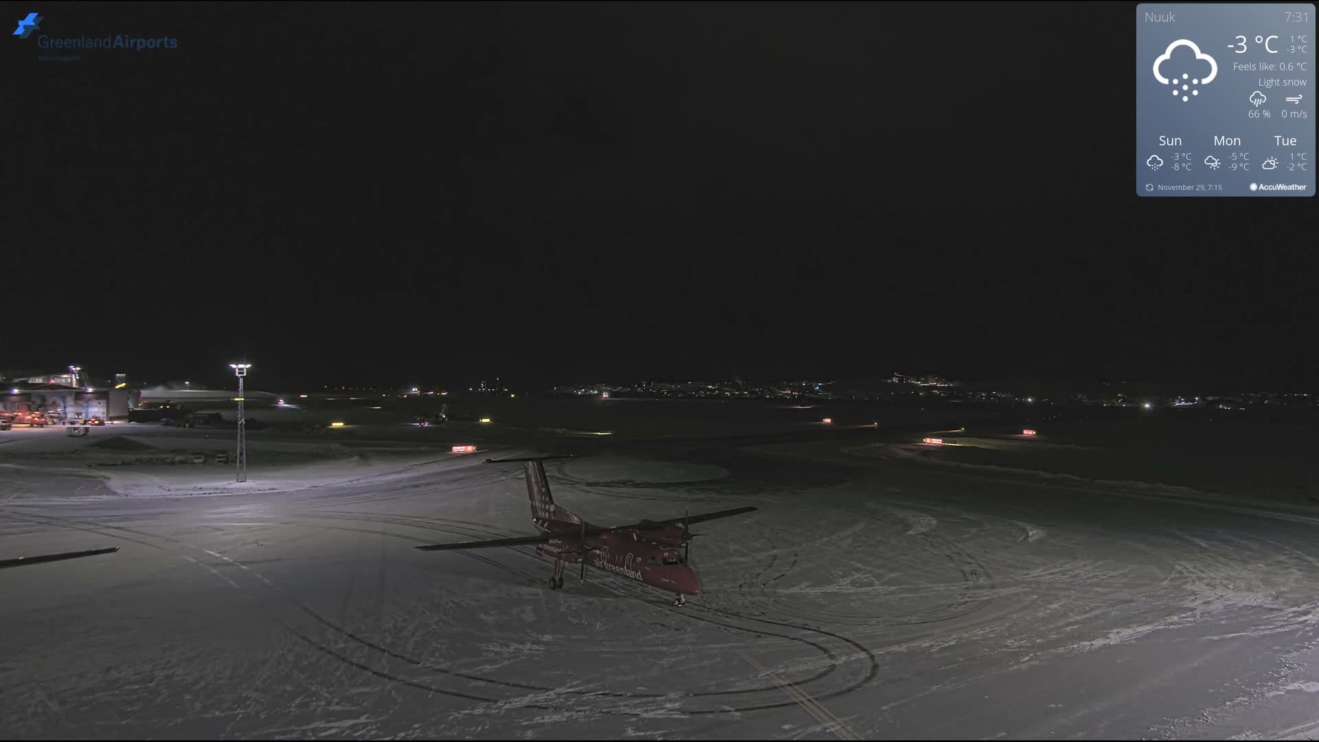 A red propeller aircraft is parked on a snow-covered airport tarmac at night, illuminated by bright airport lights, with distant city lights visible under clear, cold conditions.
