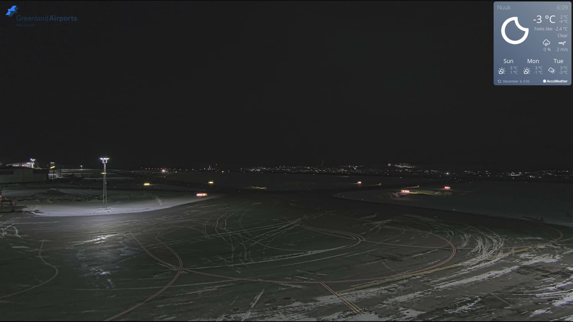 A nighttime view reveals a snow-dusted airport tarmac and runway, illuminated by ground lights with visible vehicle tracks, all under clear and cold weather conditions.