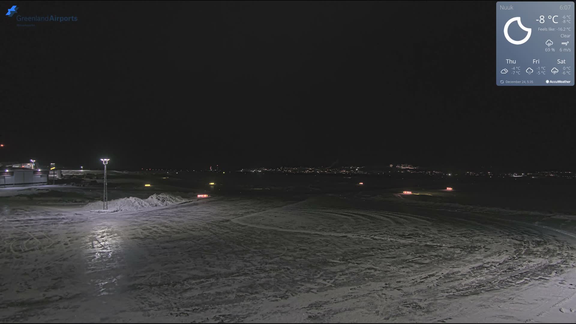 The image displays a snow and ice-covered airfield or airport tarmac at night, illuminated by various artificial lights and showing distant city lights on the horizon, under clear and cold conditions.