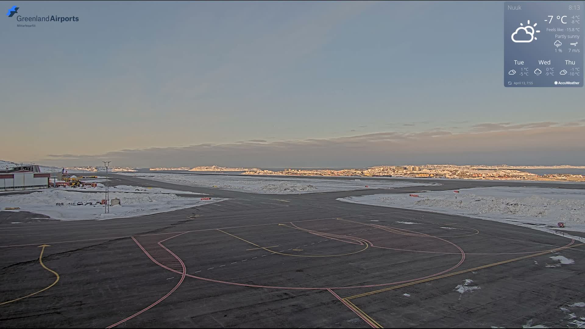 The image displays a snow and ice-covered airfield or airport tarmac at night, illuminated by various artificial lights and showing distant city lights on the horizon, under clear and cold conditions.