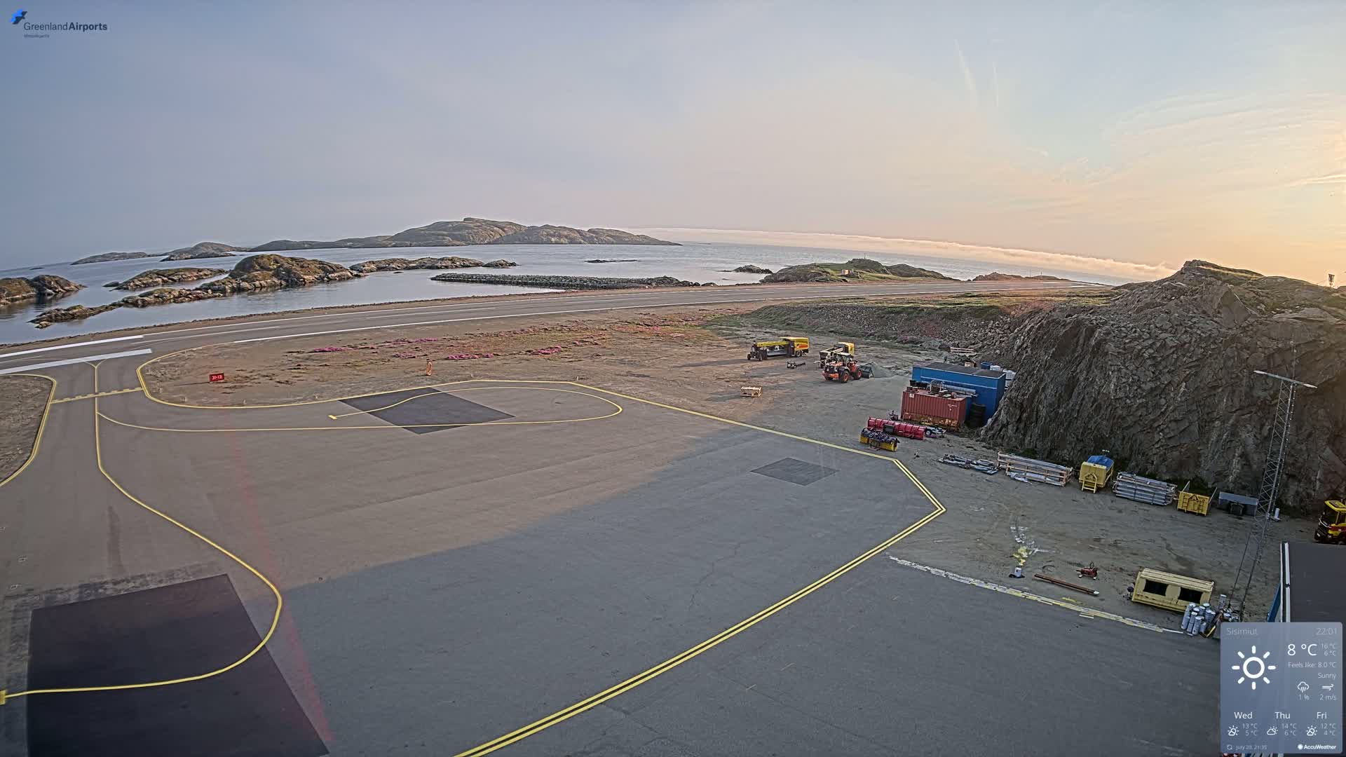 An airport tarmac with construction equipment, next to a rocky outcrop and calm ocean water with islands in the distance, under sunny skies.