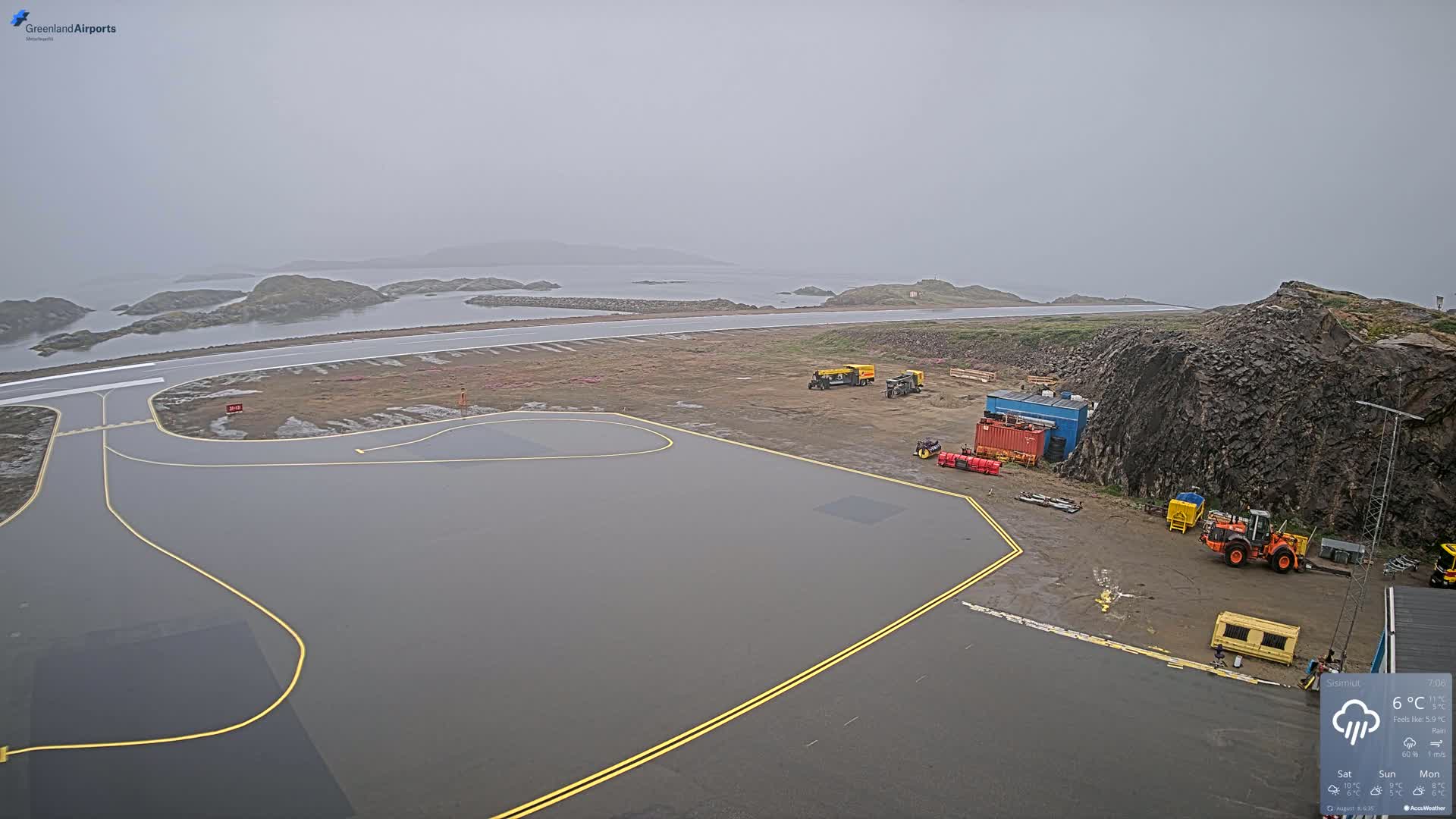 A foggy airfield with several pieces of ground maintenance equipment is situated next to a rocky shoreline and calm water.