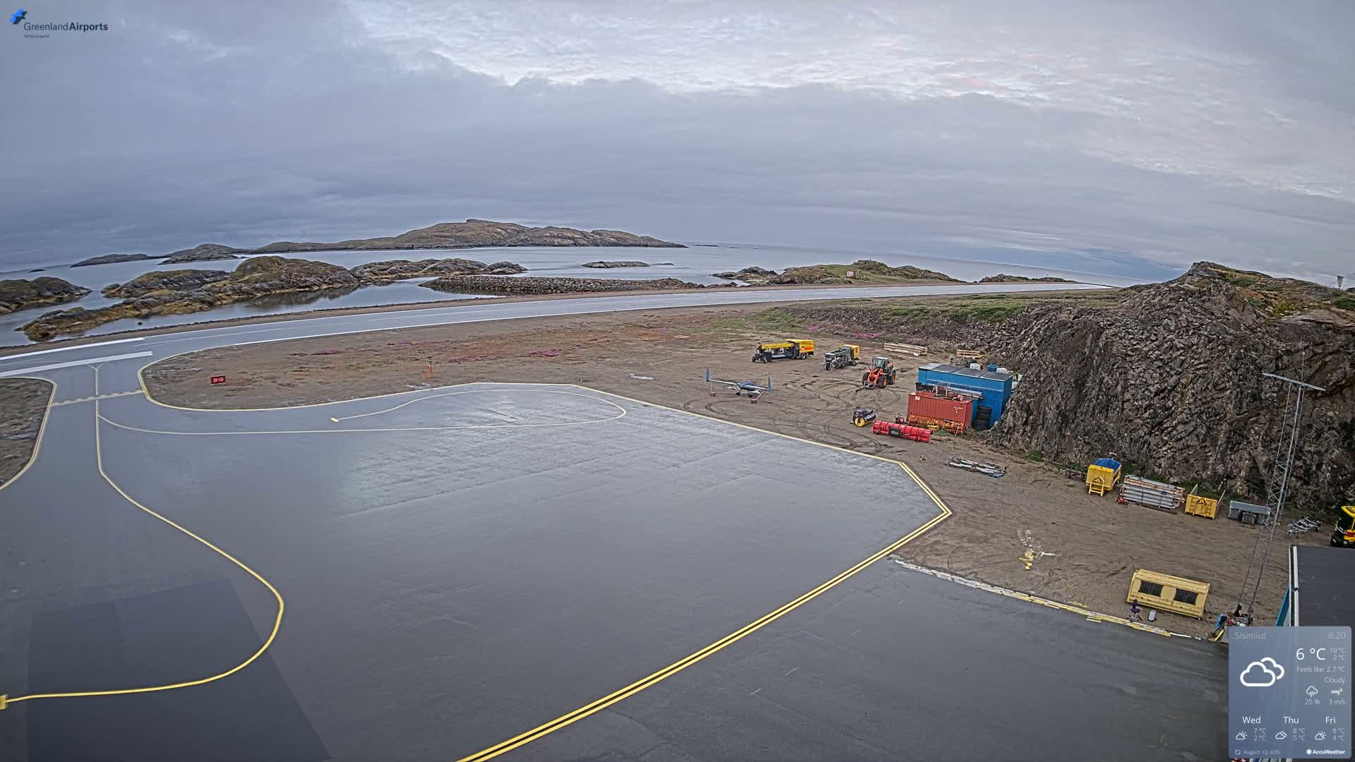 An airfield with several pieces of ground equipment sits beside a rocky coastline under a cloudy sky.