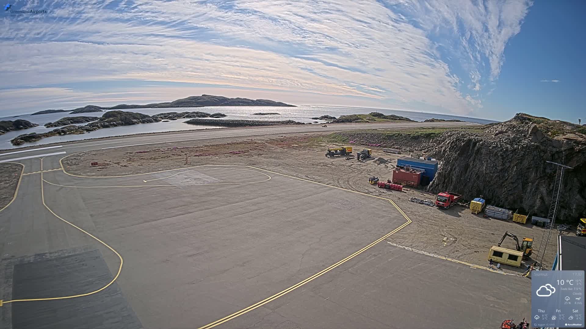 An airfield with construction equipment is visible next to a rocky coastline under a partly cloudy sky.