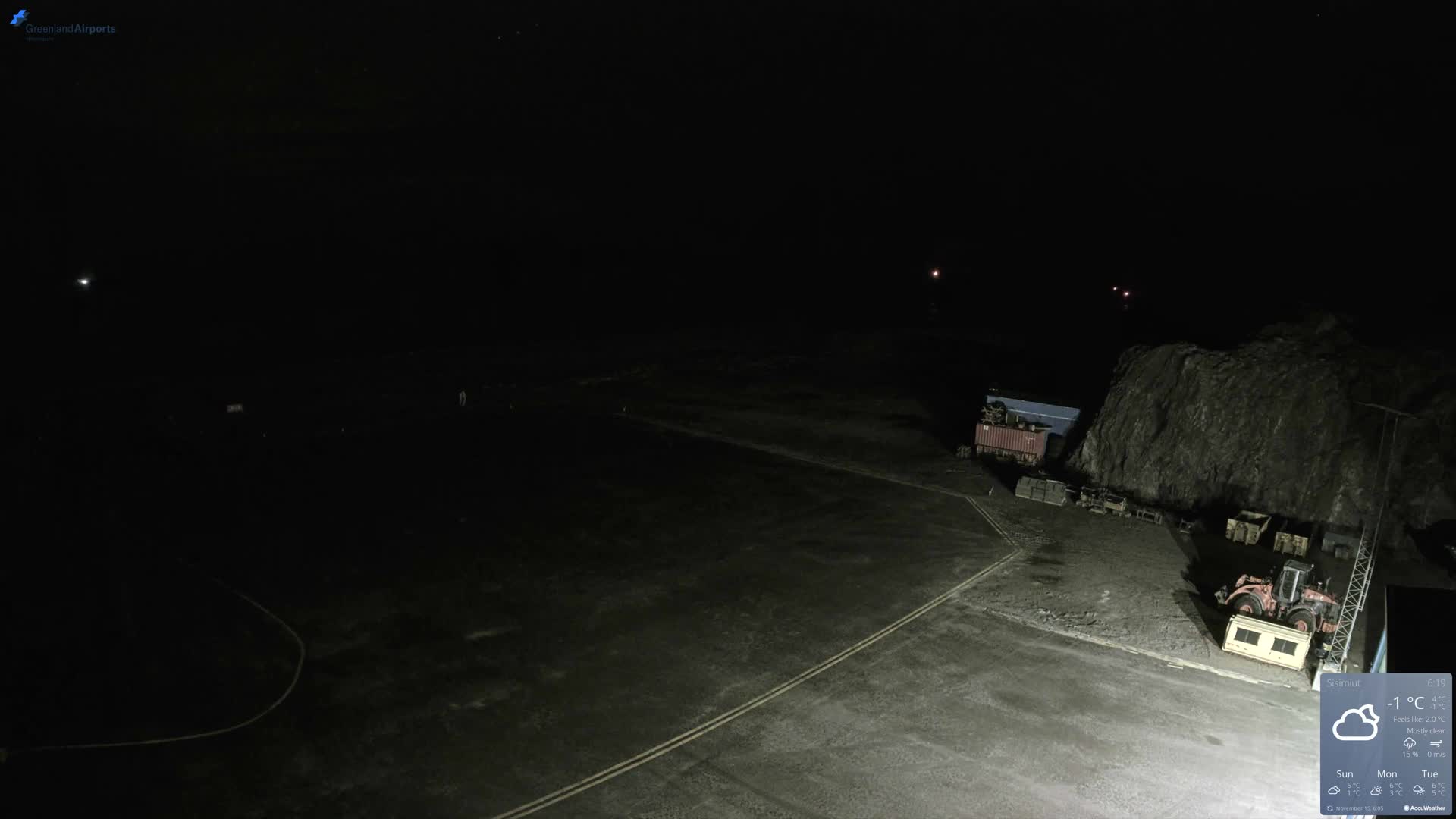 A dimly lit outdoor scene at night shows a dark paved ground with faint markings, industrial containers, and machinery against a large rocky outcrop, under a clear sky.