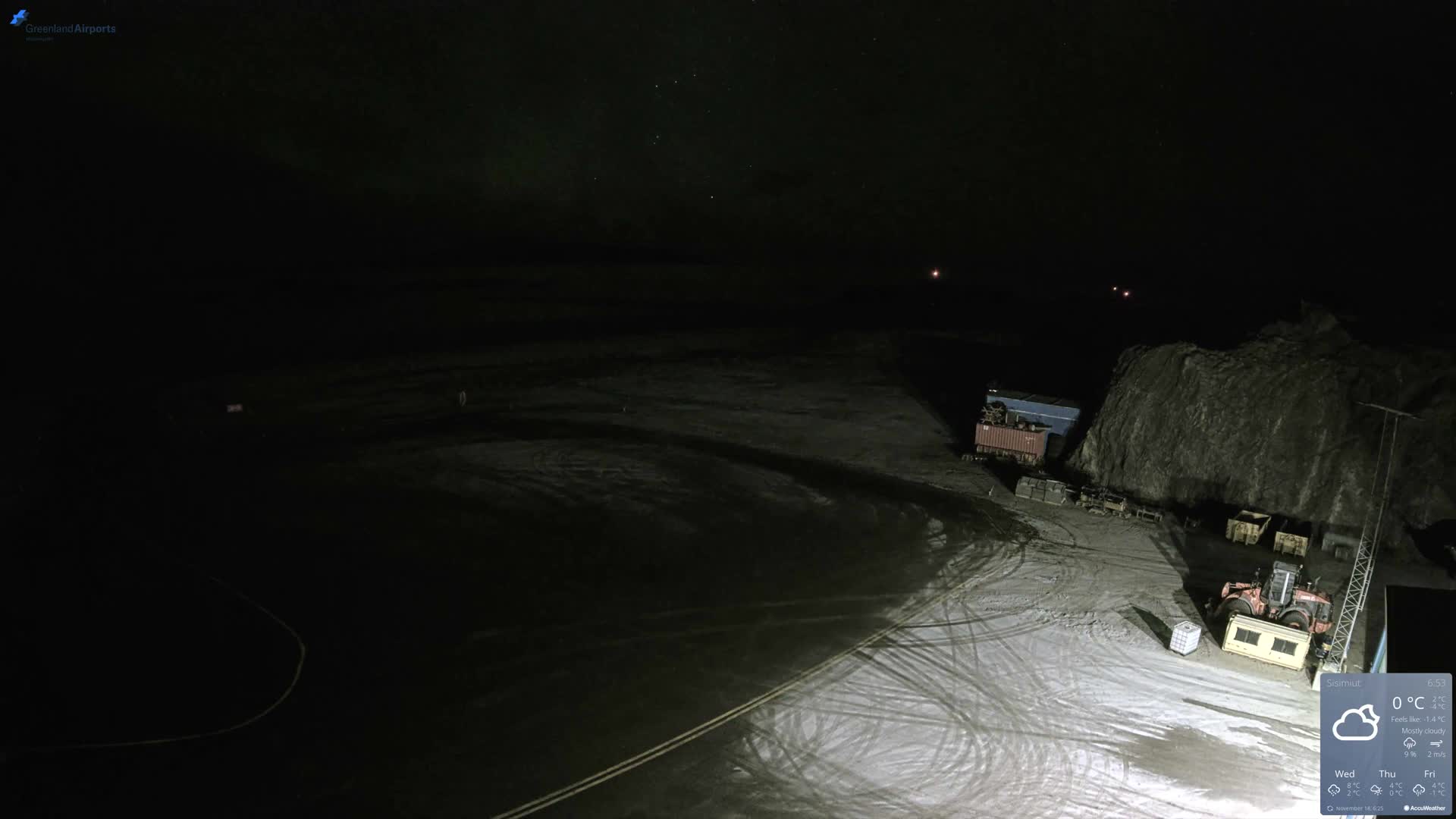 A dimly lit outdoor scene at night shows a dark paved ground with faint markings, industrial containers, and machinery against a large rocky outcrop, under a clear sky.