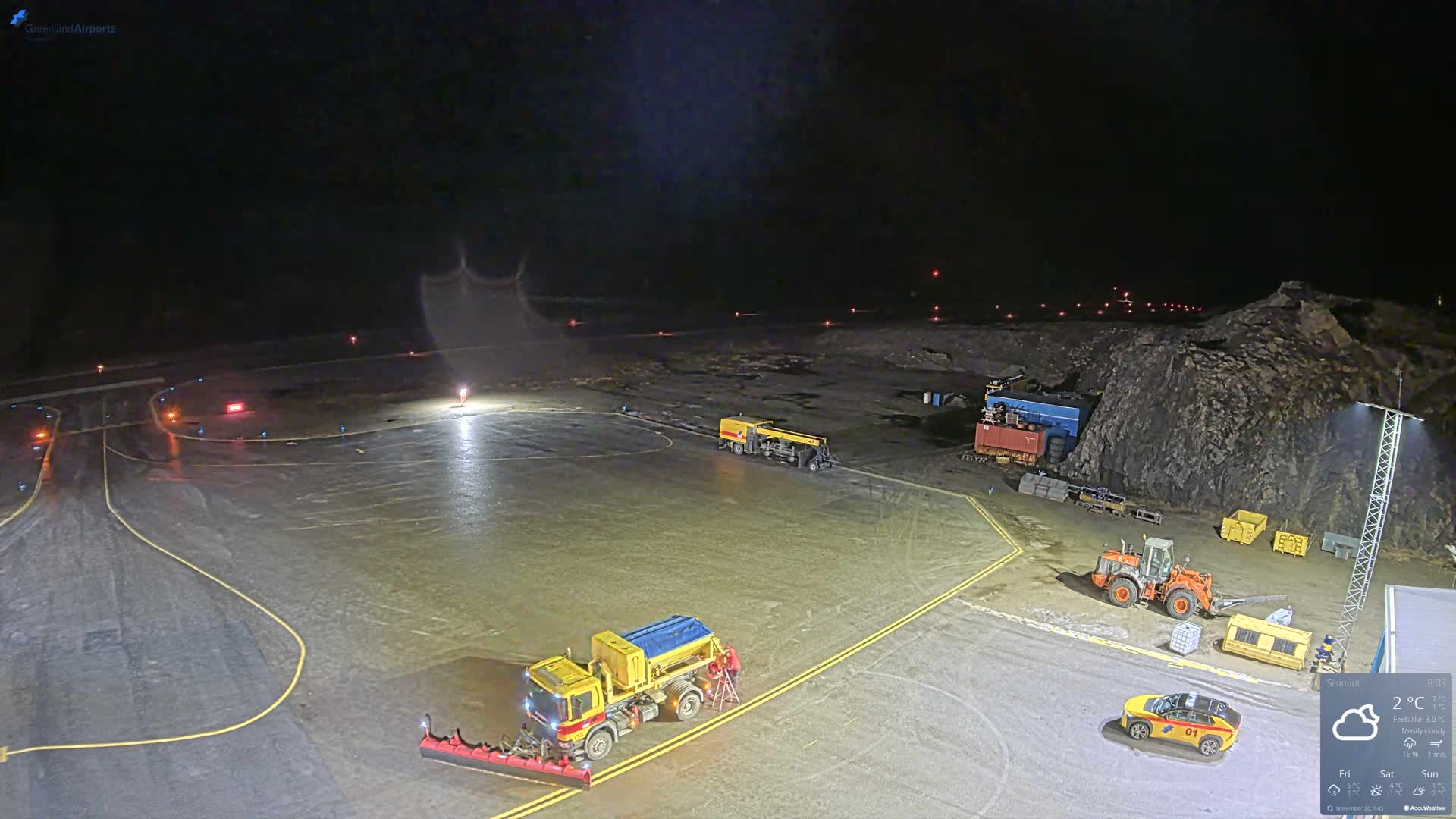 An illuminated airport tarmac is seen at night, with a large yellow snowplow truck and other maintenance vehicles parked on a wet, reflective surface beside a rocky hill.