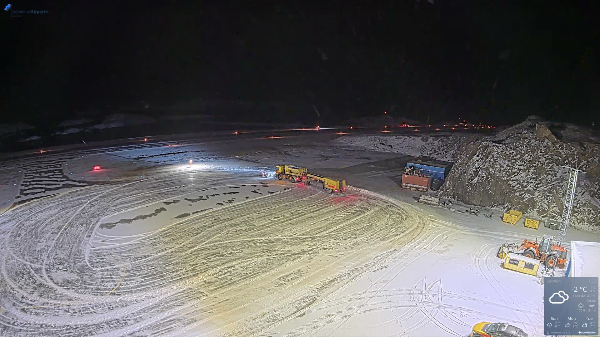 A large yellow snow-clearing vehicle operates on a snow-covered airfield at night, illuminated by ground lights under dark, wintry conditions.