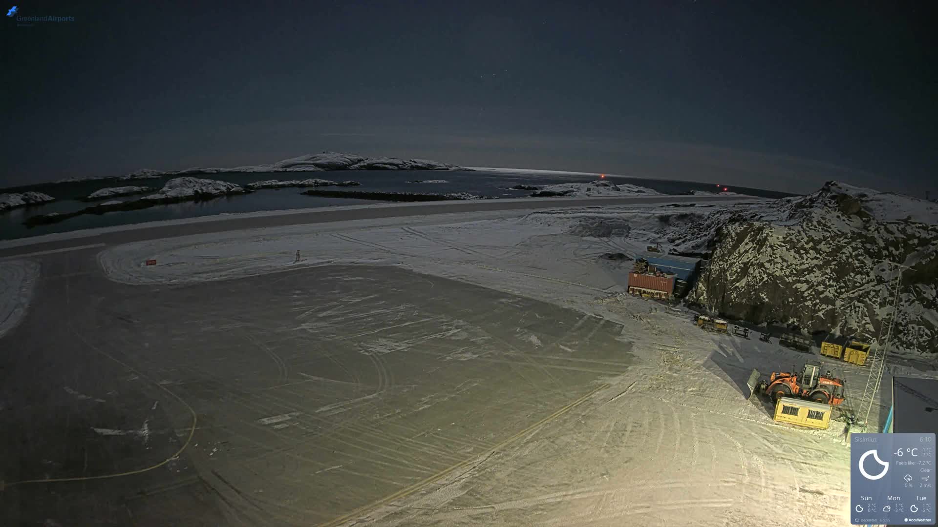 The image depicts a snowy, cold night scene at what appears to be an arctic airport or industrial site next to a body of water with islands, under a clear, star-filled sky.