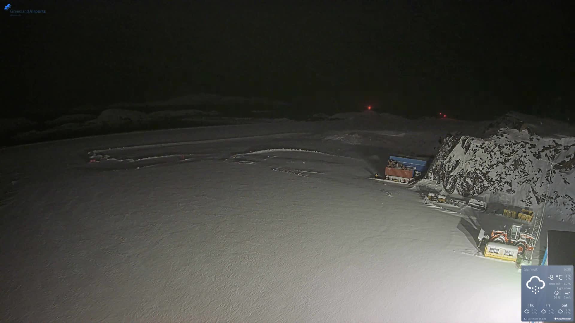 A vast, snow-covered landscape is visible at night under light snowfall and cold temperatures, featuring distant red marker lights across the plain, and a snowy rocky mountain on the right with containers and heavy machinery at its base.