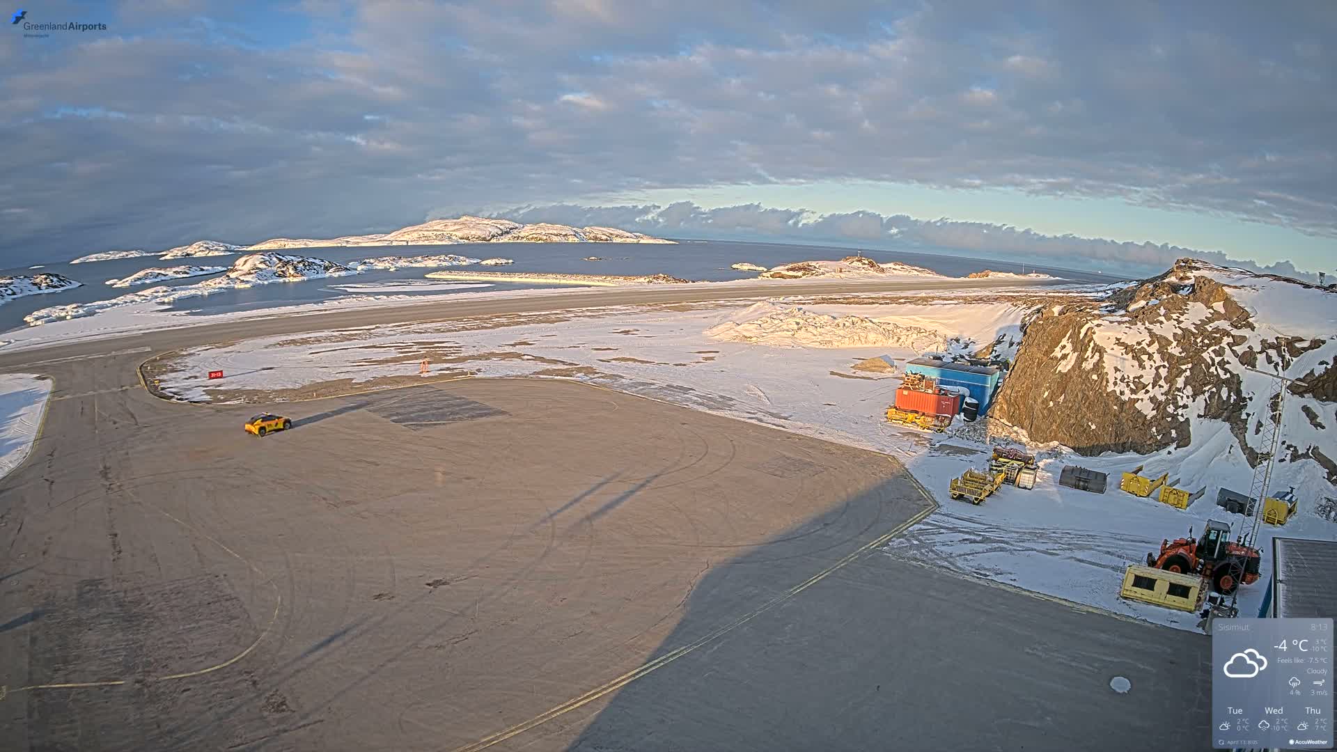 A vast, snow-covered landscape is visible at night under light snowfall and cold temperatures, featuring distant red marker lights across the plain, and a snowy rocky mountain on the right with containers and heavy machinery at its base.