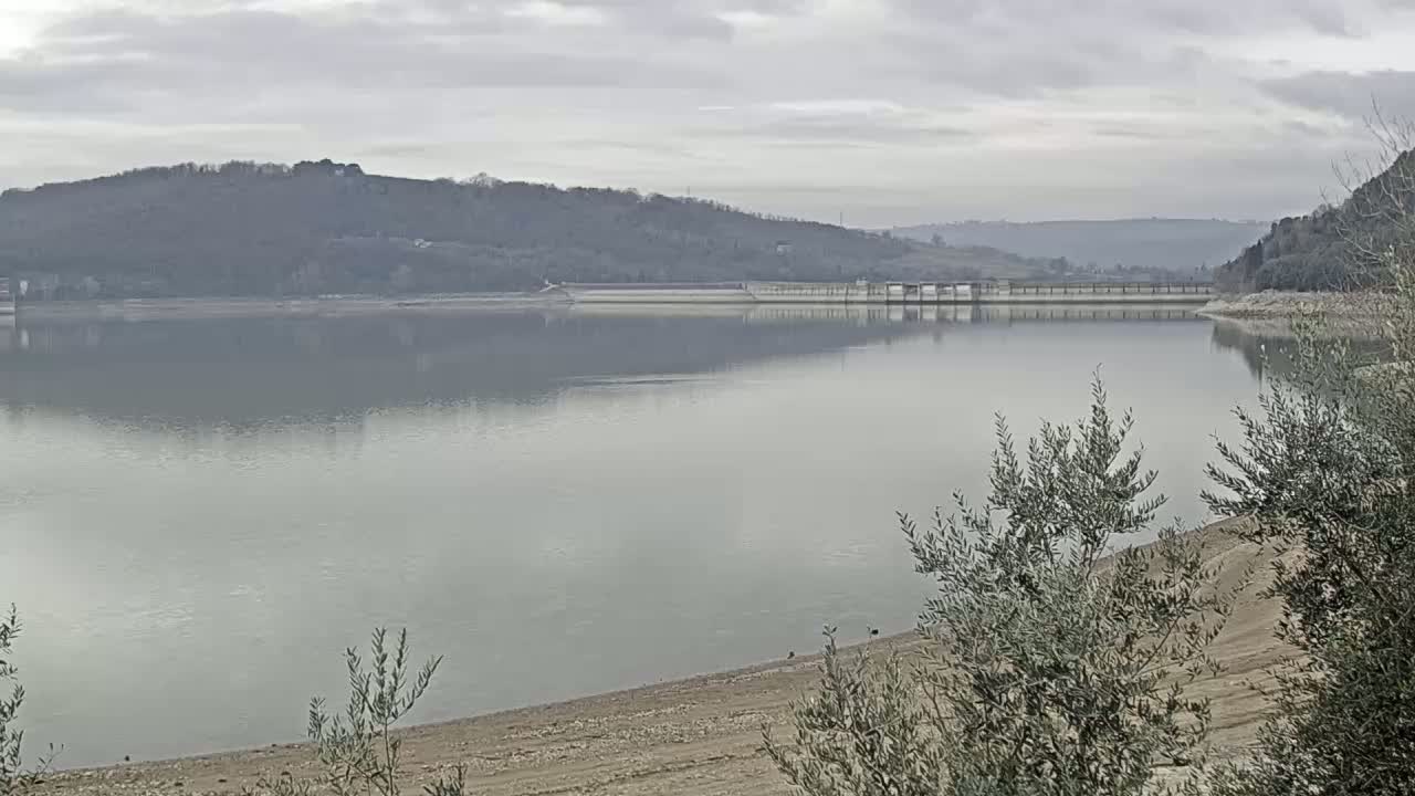 A calm lake reflects a cloudy sky, with a dam visible in the distance and low-lying vegetation in the foreground.