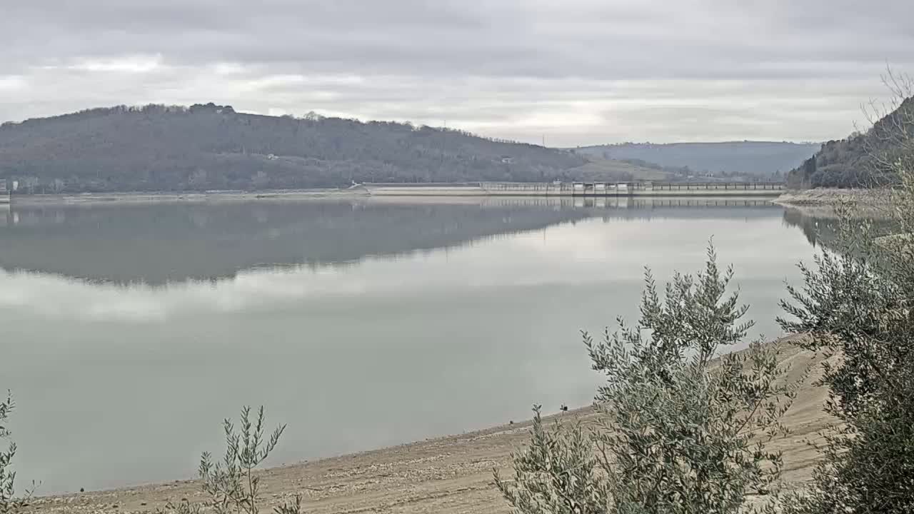 A calm lake reflecting a gray sky and hills under an overcast sky, with a dam visible in the mid-ground and exposed lakebed in the foreground.
